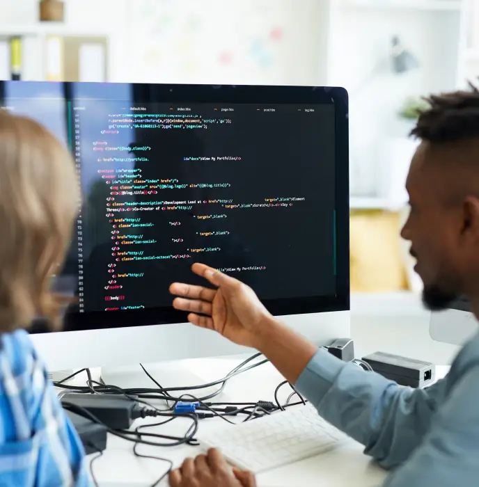 Two people sit at a desk looking at a large computer monitor displaying lines of colorful HTML and CSS code. One person gestures toward the screen while discussing the code, with cables, a keyboard, and other devices on the desk in front of them.