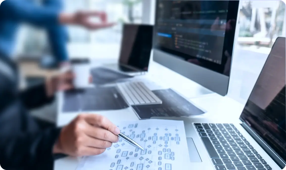 <p>A close-up of a person working at a bright, modern desk with multiple computers. One hand holds a pen while pointing at a printed flowchart, and several monitors display code. Another person gestures in the blurred background, suggesting collaboration or discussion in a software development setting.</p>