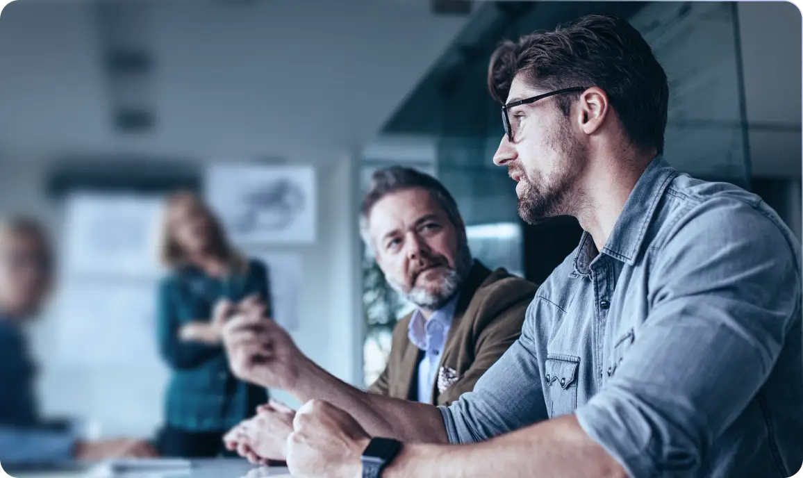 <p>A group of people in a modern office setting, with a man in the foreground speaking while another man beside him listens attentively; two blurred colleagues and whiteboards are visible in the background.</p>