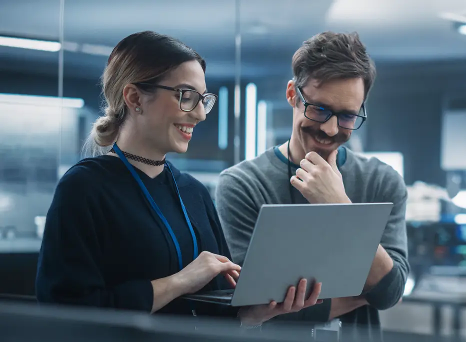 Two smiling colleagues wearing glasses collaborate while looking at a laptop in a modern office setting.