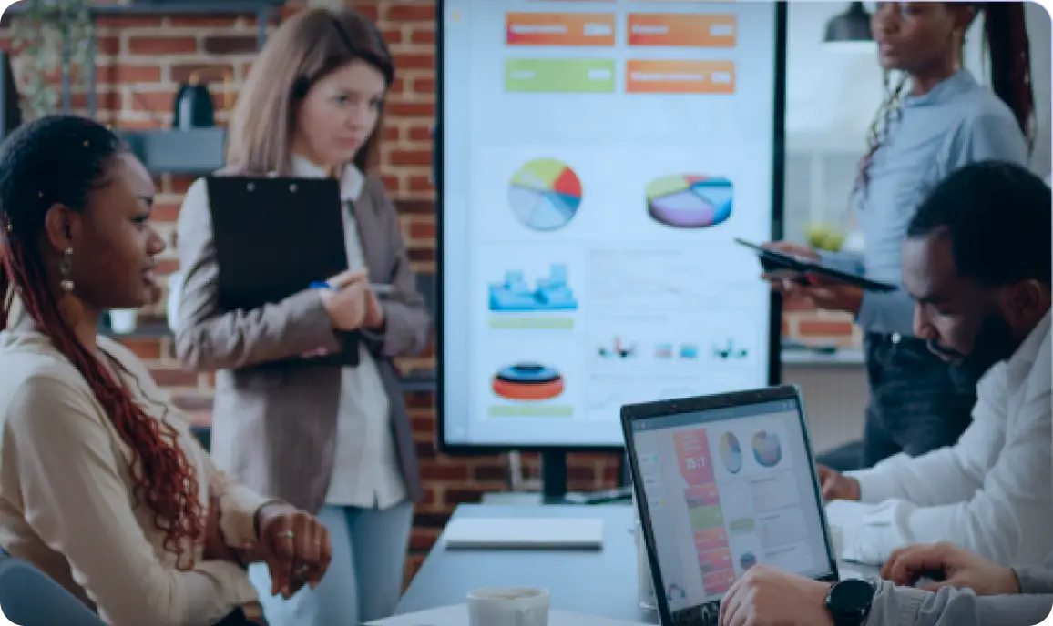 <p>A group of professionals in a modern office collaborate during a meeting; two people sit at a table working on a laptop displaying colorful charts, while two others stand near a large screen showing pie charts and data visualizations.</p>