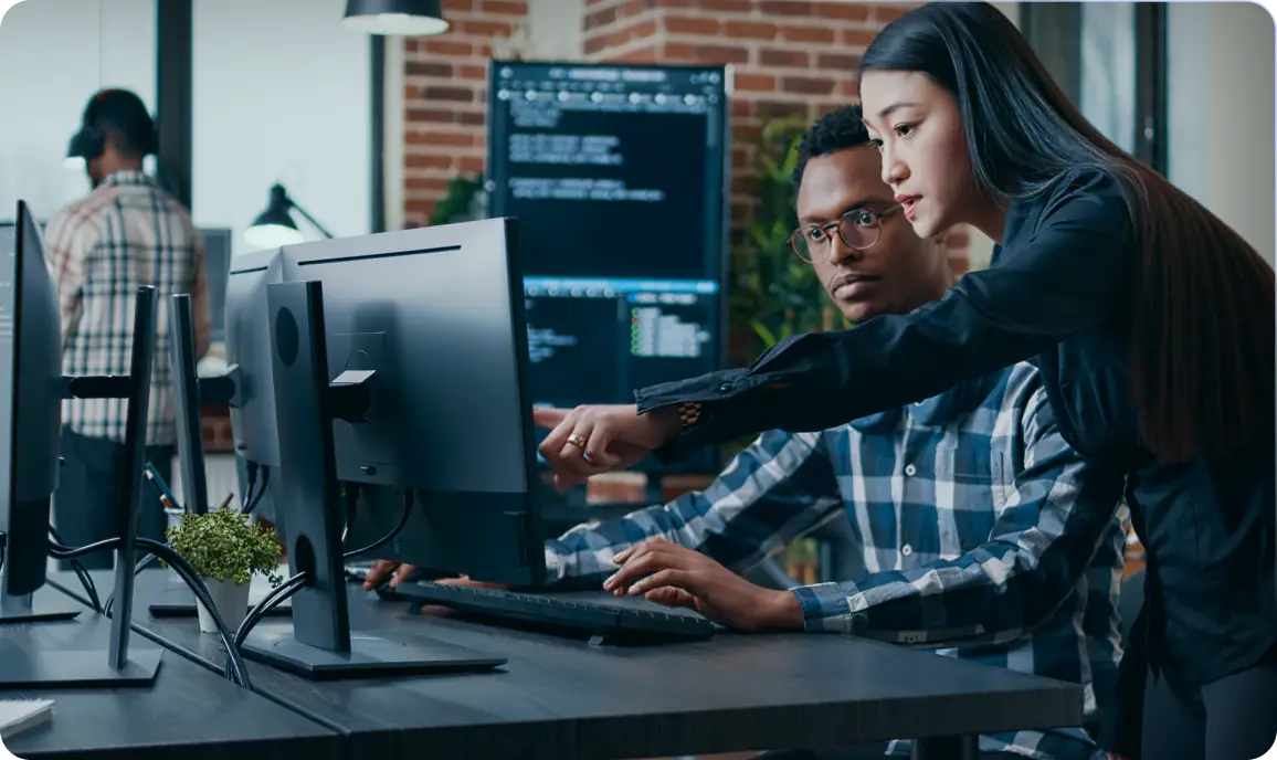 <p>A woman standing beside a seated man points at a computer monitor, guiding him as he types on the keyboard in a modern office with multiple monitors and a colleague working in the background.</p>