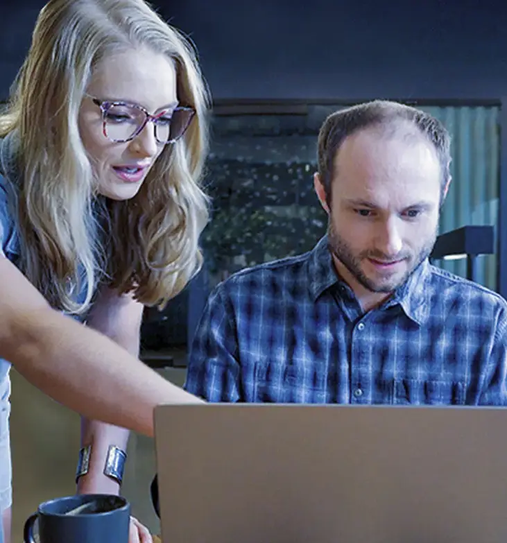 Two people collaborate at a laptop; one person is seated and focused on the screen while the other leans in and points, explaining something.
