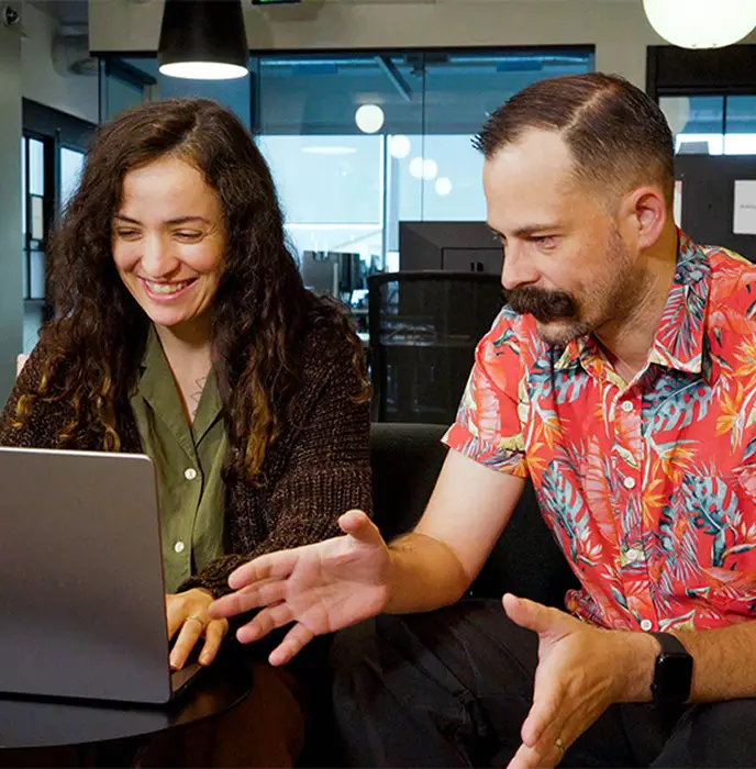 <p>Two people sitting together in an office, smiling and looking at a laptop screen while discussing something.</p>
