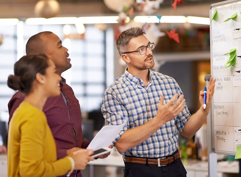 A group of three colleagues stand near a whiteboard in an office, with one person presenting and pointing to notes while the others listen and look on.
