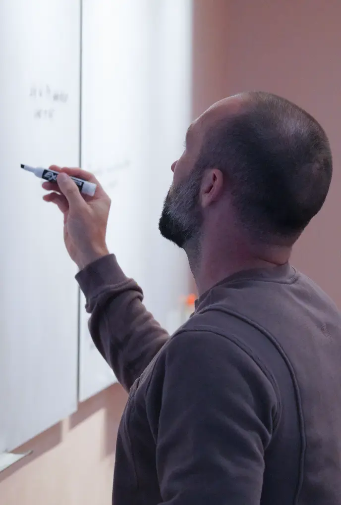 <p>A man with a shaved head and short beard writes on a whiteboard using a black marker, viewed from behind and slightly to the side. He is wearing a long-sleeve sweatshirt and appears to be focused on the writing surface.</p>