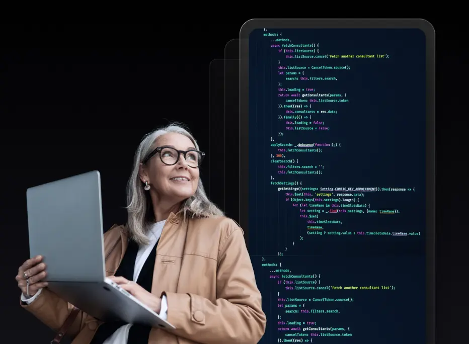 A smiling woman with long gray hair and glasses holds an open laptop while looking upward. She is wearing a tan jacket over a white shirt. Behind her, large semi-transparent screens display lines of colorful programming code on a dark background.