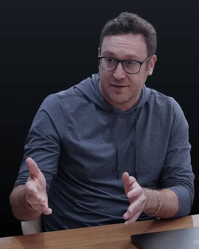 <p>A man wearing glasses and a grey hoodie sits at a table, gesturing with both hands as he speaks. A laptop is partially visible on the table in front of him, and the background is dark and blurred.</p>