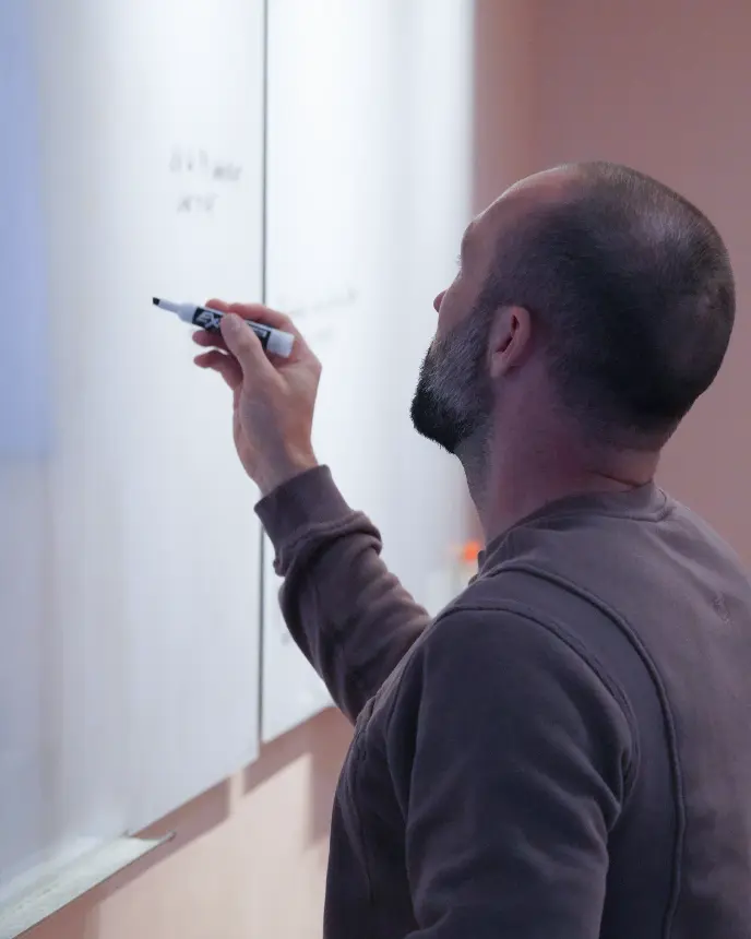 A man with a shaved head and beard writes on a large whiteboard using a black dry-erase marker. He is viewed from behind and appears to be focused on the notes he is adding to the board.