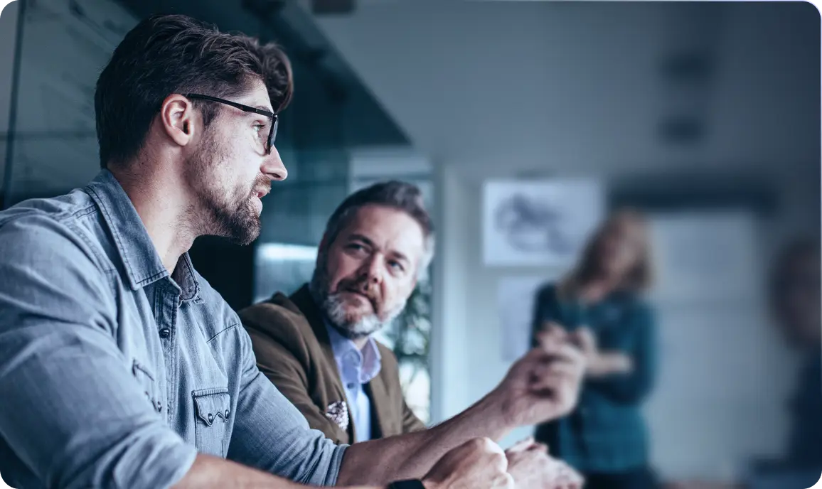 <p>Two men sitting in a meeting room, engaged in conversation; one with glasses and a beard speaking, while the other listens attentively. A blurred person stands in the background near a whiteboard.</p>