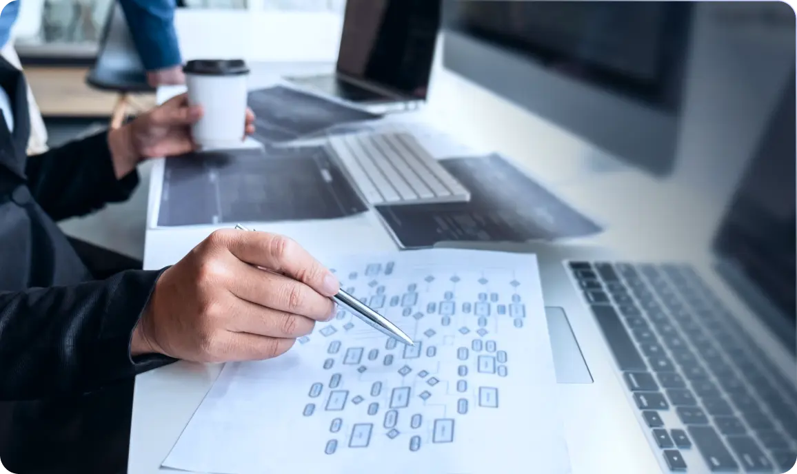 <p>A person sitting at a desk points to a printed flowchart with a pen, surrounded by a laptop, keyboard, and documents. Another person in the background holds a coffee cup. The scene suggests planning, analysis, or software/system design work.</p>