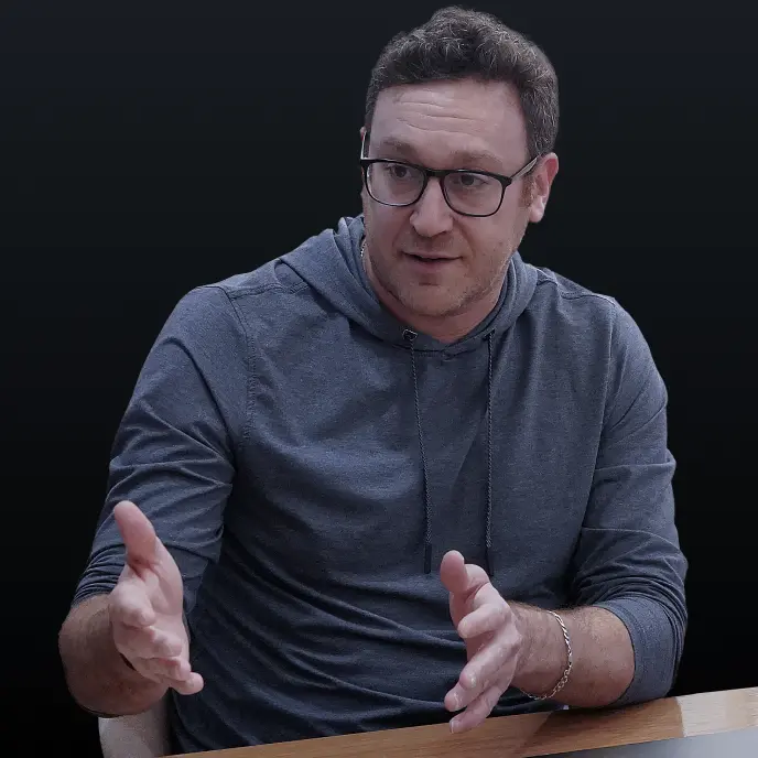 <p>A man with short curly hair and glasses sits at a table, gesturing with his hands as he speaks. He is wearing a gray hoodie and appears to be engaged in a discussion against a dark background.</p>