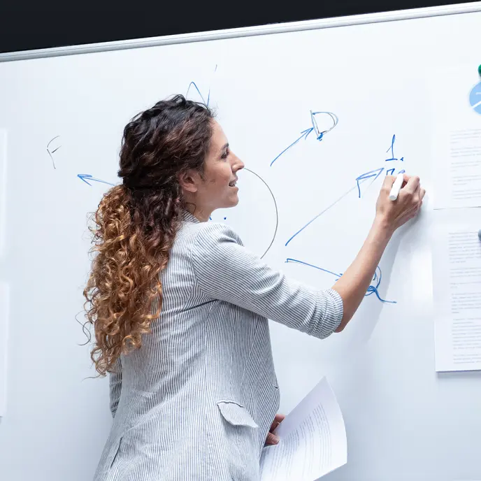 <p>A woman with curly hair stands in front of a whiteboard, writing diagrams and arrows with a marker while holding documents in her other hand. She appears to be explaining or mapping out a process.</p>