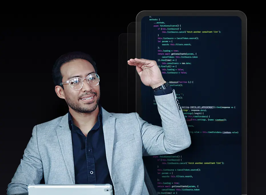 A man wearing glasses and a light gray blazer gestures with one hand raised while sitting in front of a tablet, with lines of code displayed on large digital screens behind him.