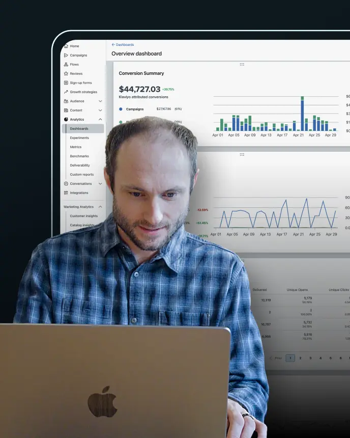 A man wearing a blue plaid shirt is working on a laptop with an Apple logo, while behind him a bright digital analytics dashboard is displayed. The dashboard shows graphs, charts, and conversion summary data, including bar and line charts, representing performance metrics and trends.
