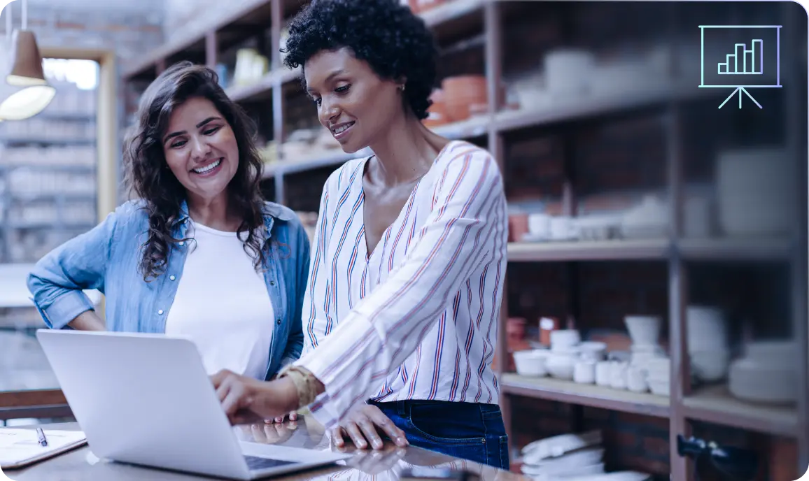 <p>Two women stand together in a shop or studio space, smiling as they look at a laptop screen. Shelves filled with pottery or ceramic items are visible in the background. One woman points at the laptop while the other watches happily. A small presentation or bar-chart icon appears in the upper-right corner, suggesting growth or business analytics.</p>