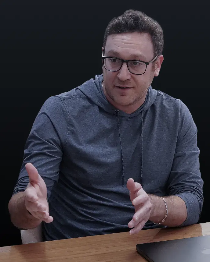 <p>A man wearing glasses and a grey hoodie sits at a wooden table, gesturing with both hands as he speaks. A closed laptop rests on the table in front of him against a dark background.</p>
