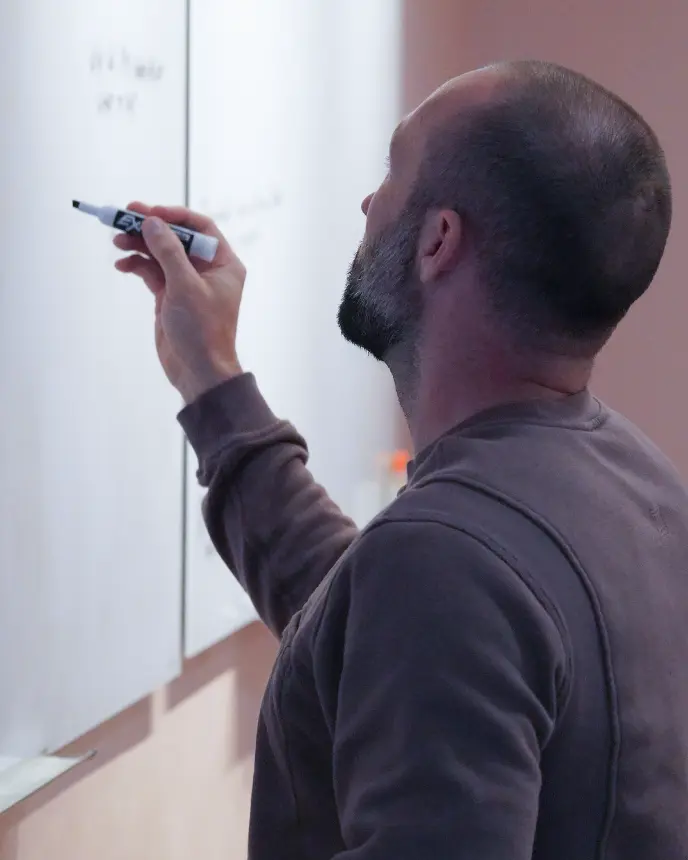 <p>A man seen from behind writes on a whiteboard with a marker, focusing intently as he adds notes to the board.</p>