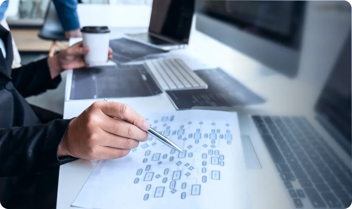 <p>A person sitting at a desk points to a printed flowchart with a pen, while holding a coffee cup in the other hand. Computer screens, a keyboard, and documents are spread across the workspace, indicating planning, analysis, or software project design.</p>