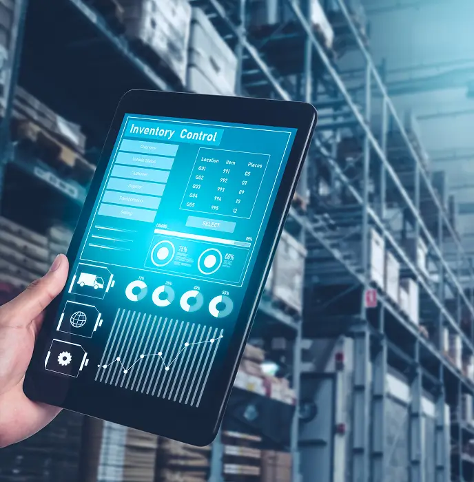 <p>A person holding a tablet displaying an &ldquo;Inventory Control&rdquo; dashboard with charts, graphs, and item data, while standing inside a large warehouse filled with shelves and stacked boxes.</p>