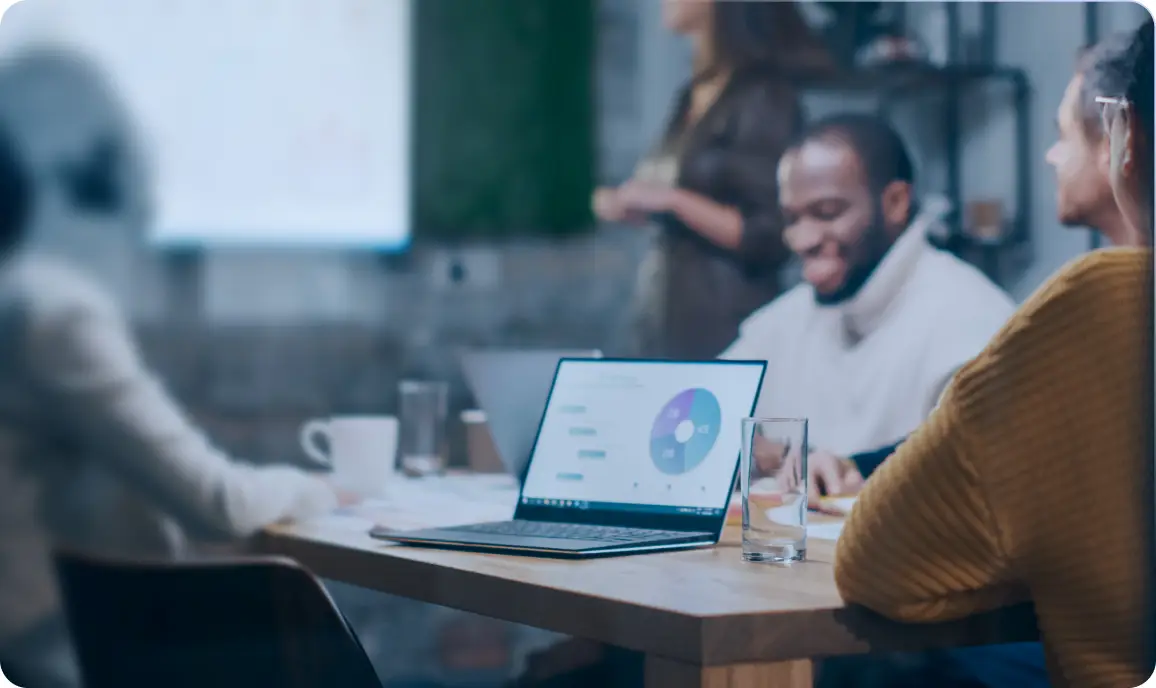 <p>Several people sit around a wooden table in a modern meeting room. A laptop displaying a colorful chart is open in the center of the table, surrounded by papers, glasses of water, and a coffee mug. The group appears to be engaged in discussion, with one person standing near a screen in the background and others seated, slightly out of focus.</p>