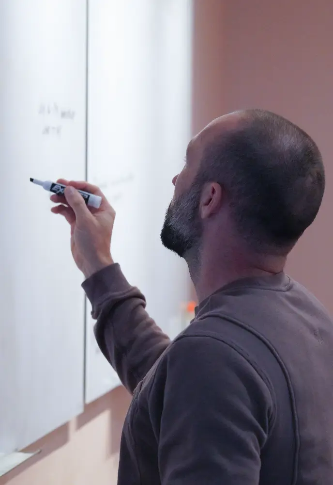 <p>A person writing on a whiteboard with a marker, shown from the side and slightly behind, focusing on their hand and the board.</p>
