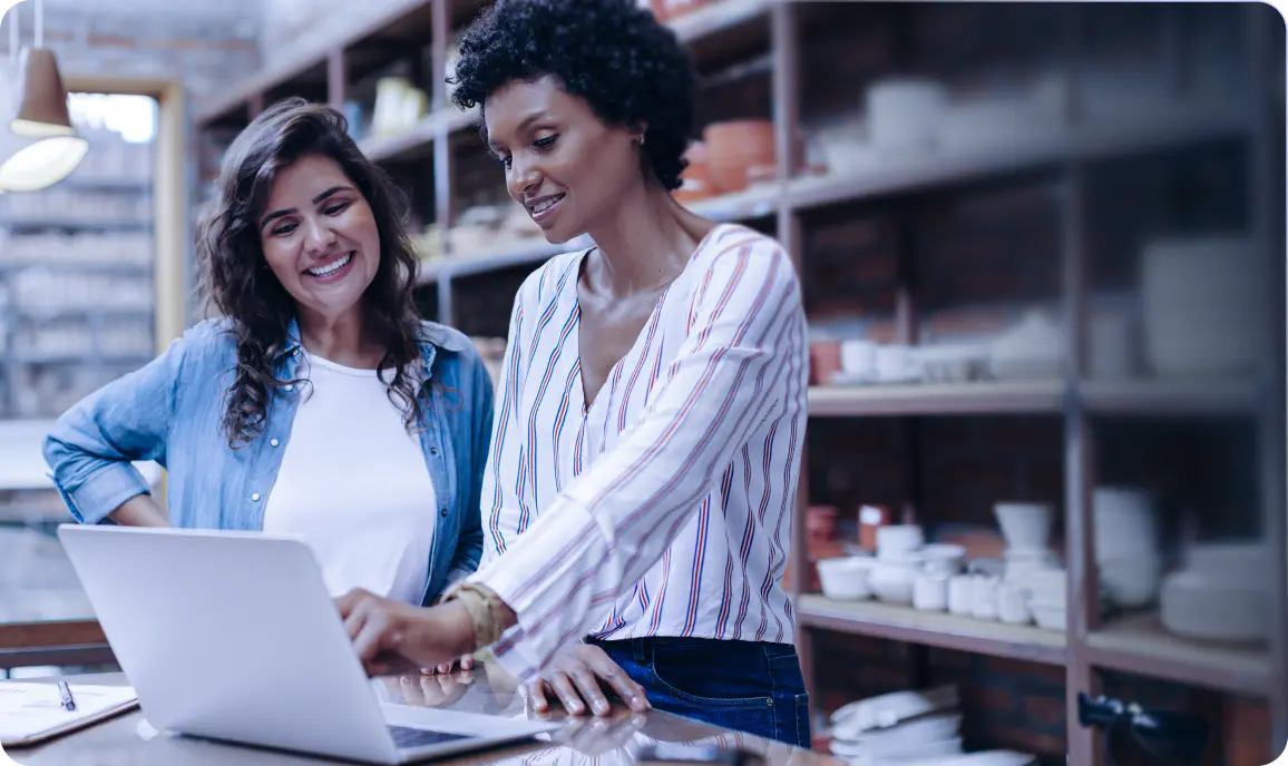 <p>Two women standing together in a shop or studio, smiling as they look at a laptop screen. One woman is pointing at the laptop while the other stands beside her. Shelves filled with pottery and home goods are visible in the background.</p>