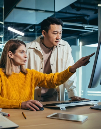 <p>Two coworkers in a modern office discuss content on a desktop computer screen; the woman in a yellow sweater points at the monitor while the man in a white jacket leans in attentively.</p>