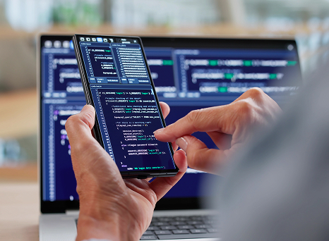 Close-up of a person using a smartphone to write or debug code, with a laptop displaying code in the background, suggesting software development or mobile-first programming.