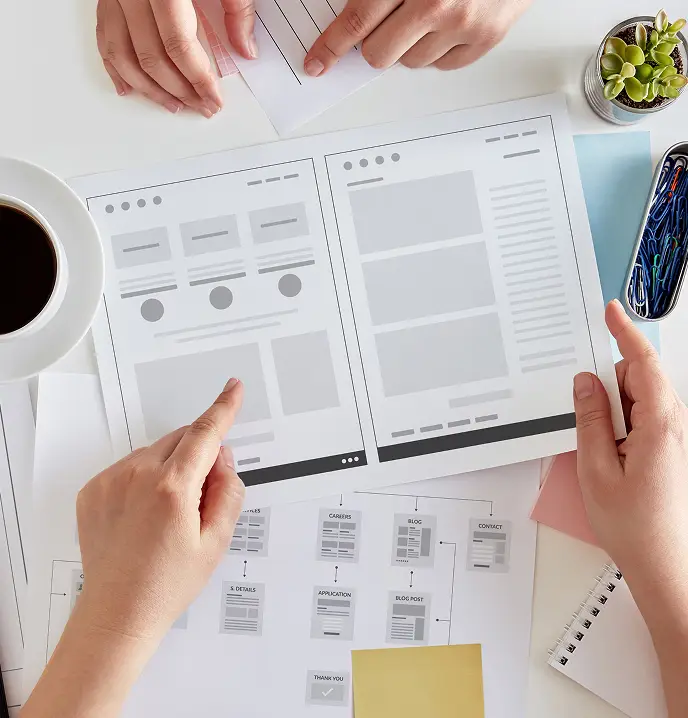 <p>Hands of several people reviewing printed website wireframes and a sitemap diagram on a desk, surrounded by office supplies including a cup of coffee, sticky notes, and a small potted plant.</p>