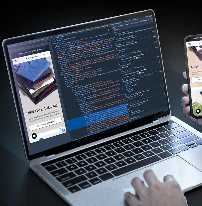 <p>Laptop displaying website code and a mobile-responsive preview side-by-side, with a person&rsquo;s hands using the laptop while holding a smartphone showing the same website layout.</p>