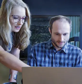 A woman with long blonde hair and glasses is leaning over and pointing at a laptop screen while explaining something to a man in a blue plaid shirt who is attentively looking at the screen.