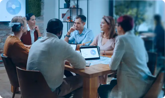 <p>Group of professionals engaged in a meeting around a wooden table, with laptops displaying charts, suggesting a data-driven team discussion or strategic planning session in a modern office setting.</p>