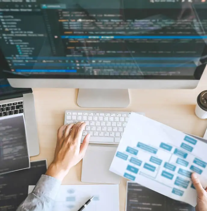 <p>A person working at a desk with a large desktop monitor displaying code, holding a printed system diagram in one hand while typing on a keyboard with the other. Laptops and documents surround the workspace, suggesting a development or technical planning session.</p>