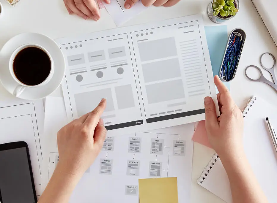 Two people reviewing website wireframe designs printed on paper, surrounded by office supplies including a cup of coffee, smartphone, and a layout flow diagram on a white desk.