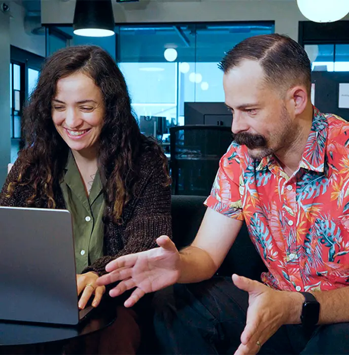 <p>Two people sitting together in a modern office space, smiling and looking at a laptop screen. The woman on the left is typing while the man on the right gestures with his hands as they discuss something on the laptop.</p>