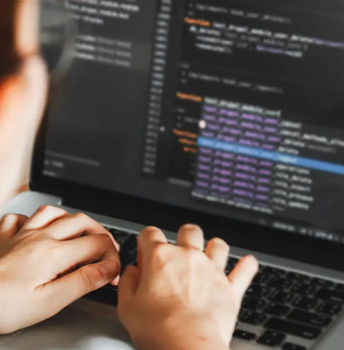 <p>Close-up view of a person typing on a laptop with a dark-themed code editor open on the screen, showing lines of code highlighted in various colors.</p>