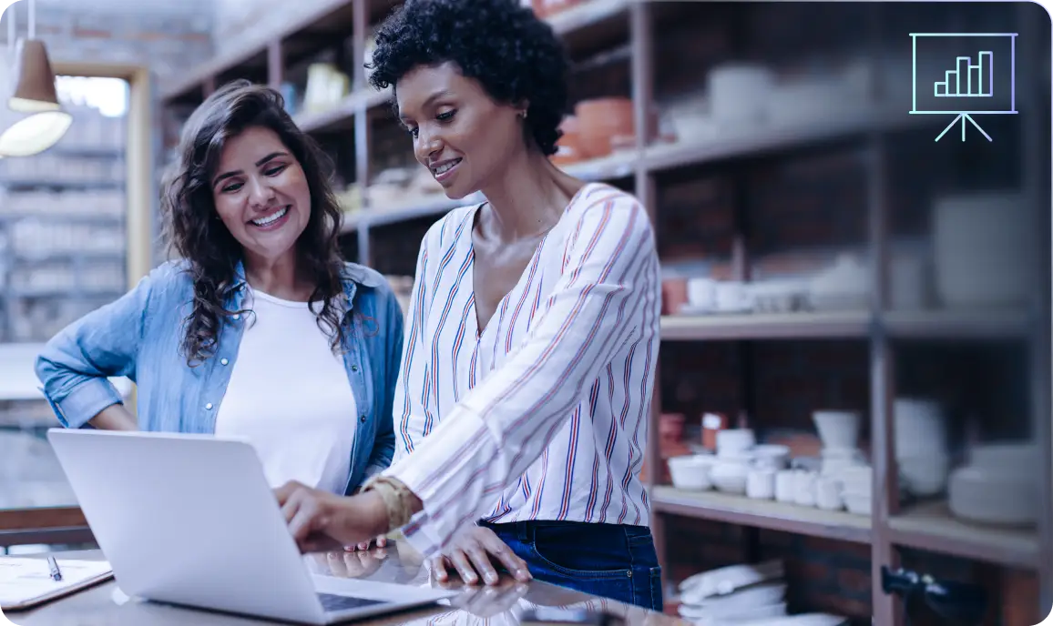 <p>Two women standing together in a shop or studio, smiling as they look at a laptop screen on the counter, with shelves of pottery and home goods in the background and a small chart icon displayed in the top-right corner.</p>