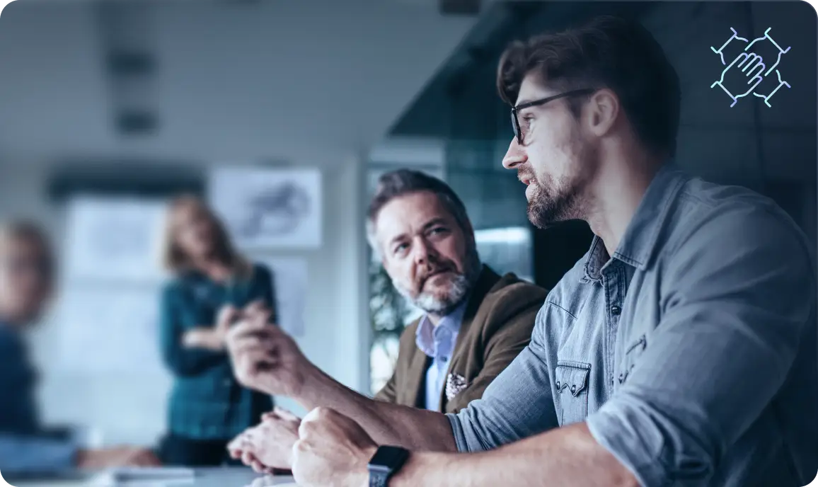 <p>Two men sitting at a meeting table engaged in conversation. One man with glasses is speaking while the other listens attentively. Blurred colleagues and office elements are visible in the background, with an icon of interconnected hands in the upper right corner.</p>