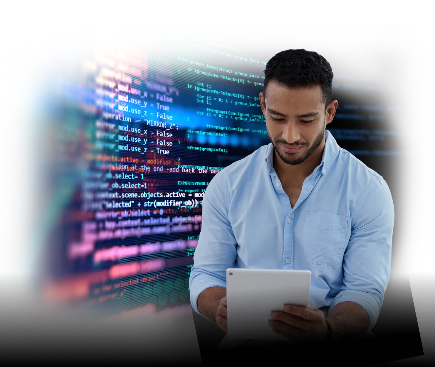 A man in a light blue shirt sits and looks down at a tablet he is holding, with a large, colorful display of computer code and digital graphics in the background.