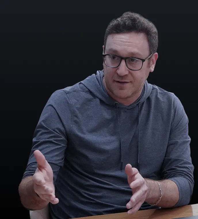 <p>A man wearing glasses and a gray hoodie sits at a table, gesturing with both hands as he speaks, against a dark background.</p>