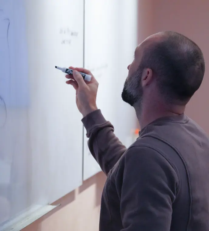 <p>A man with a shaved head and beard writes on a large whiteboard using a marker, viewed from behind, as he works on notes or diagrams in a classroom or office setting.</p>