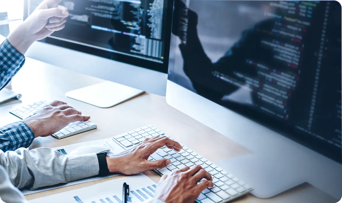 <p>A close-up view of software developers working at a desk with large monitors displaying lines of code. One person is typing on a keyboard while another is pointing at the screen, suggesting collaboration or problem-solving. Documents and office supplies are visible on the desk.</p>