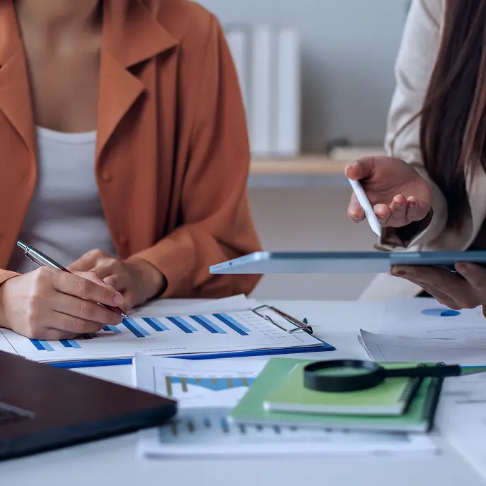 <p>Two people collaborate at a desk, reviewing printed charts and graphs while one person writes with a pen and the other uses a stylus on a tablet, suggesting data analysis, planning, or a business meeting.</p>