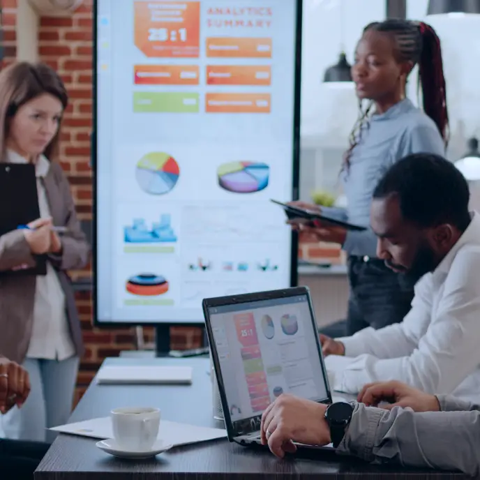 <p>A diverse team works in a modern office, reviewing analytics dashboards with charts and graphs displayed on a large screen while one person uses a laptop and others discuss results using a tablet and notes, suggesting a collaborative data or marketing meeting.</p>