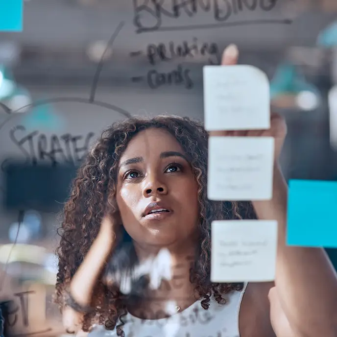 <p>A woman arranges sticky notes on a glass board covered with handwritten words like &ldquo;Strategy&rdquo; and &ldquo;Branding,&rdquo; suggesting brainstorming, planning, or creative problem-solving in a modern workspace.</p>