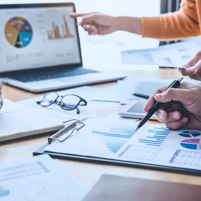 Hands point and write on printed charts and graphs spread across a desk, with a laptop displaying data visualizations in the background, suggesting collaborative data analysis or business performance review.