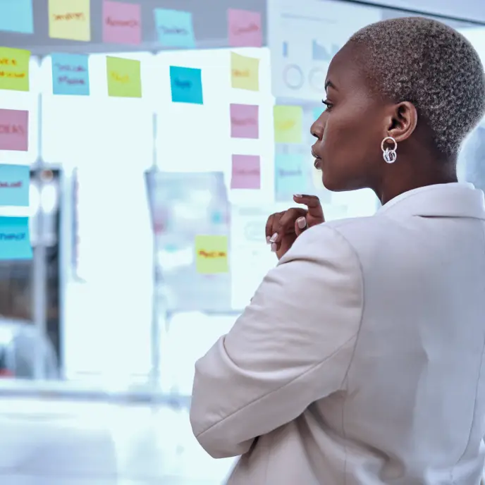 A professional woman stands in profile, thoughtfully reviewing a glass wall covered with colorful sticky notes and charts, suggesting strategic planning, brainstorming, or project management in a modern office.