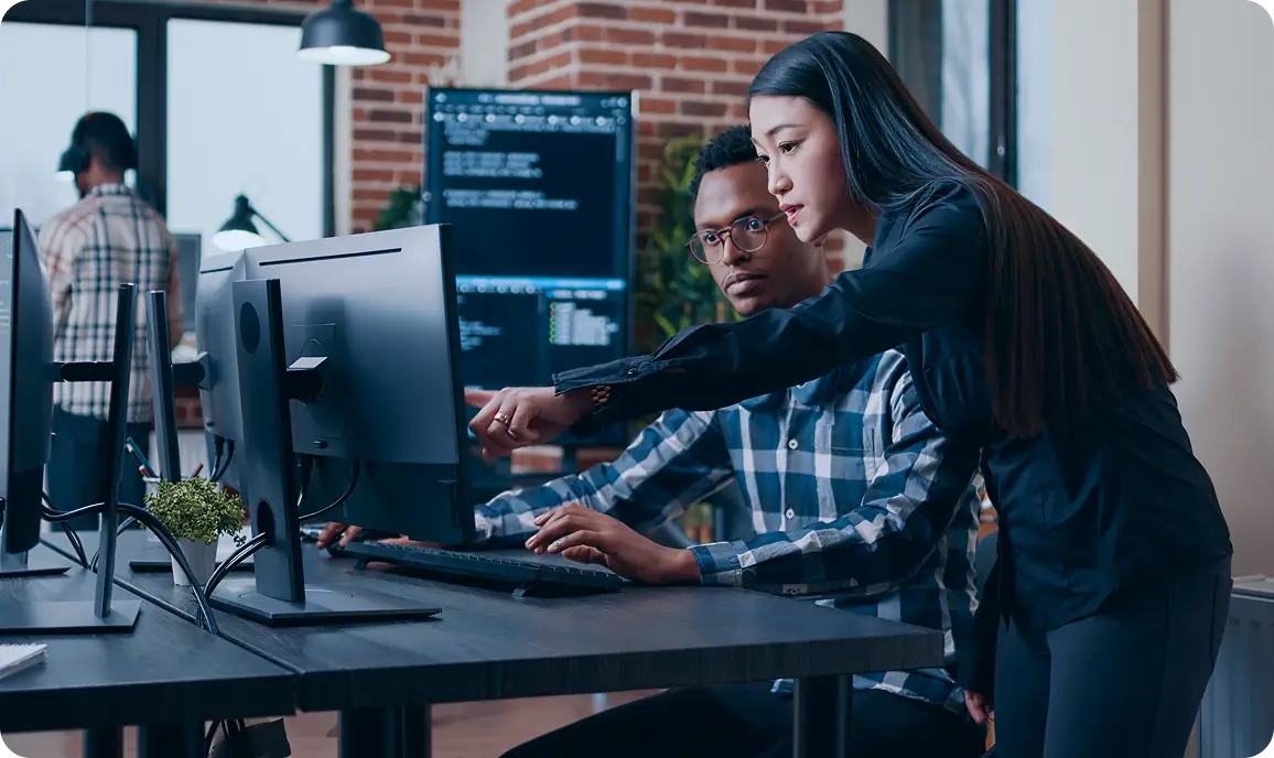 <p>A woman leans over and points at a computer monitor while a seated man types at a desk in a modern office; code is visible on screens behind them, suggesting collaboration on a software or technical task.</p>