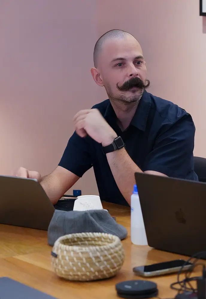 <p>A person with a shaved head and a large curled mustache sits at a wooden table, working between two open laptops. They are wearing a dark short-sleeved shirt and a smartwatch. Various items, including a small woven basket, a gray cloth, a smartphone, and a water bottle, are on the table. The background is a softly lit, pale pink wall.</p>